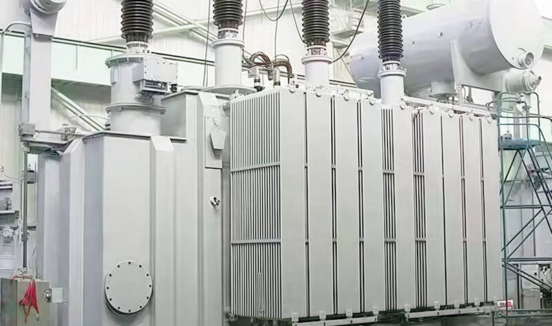 Side view of a oil immersed electrical transformer stationed inside the manufacturer's facility, emphasizing its size and quality control.